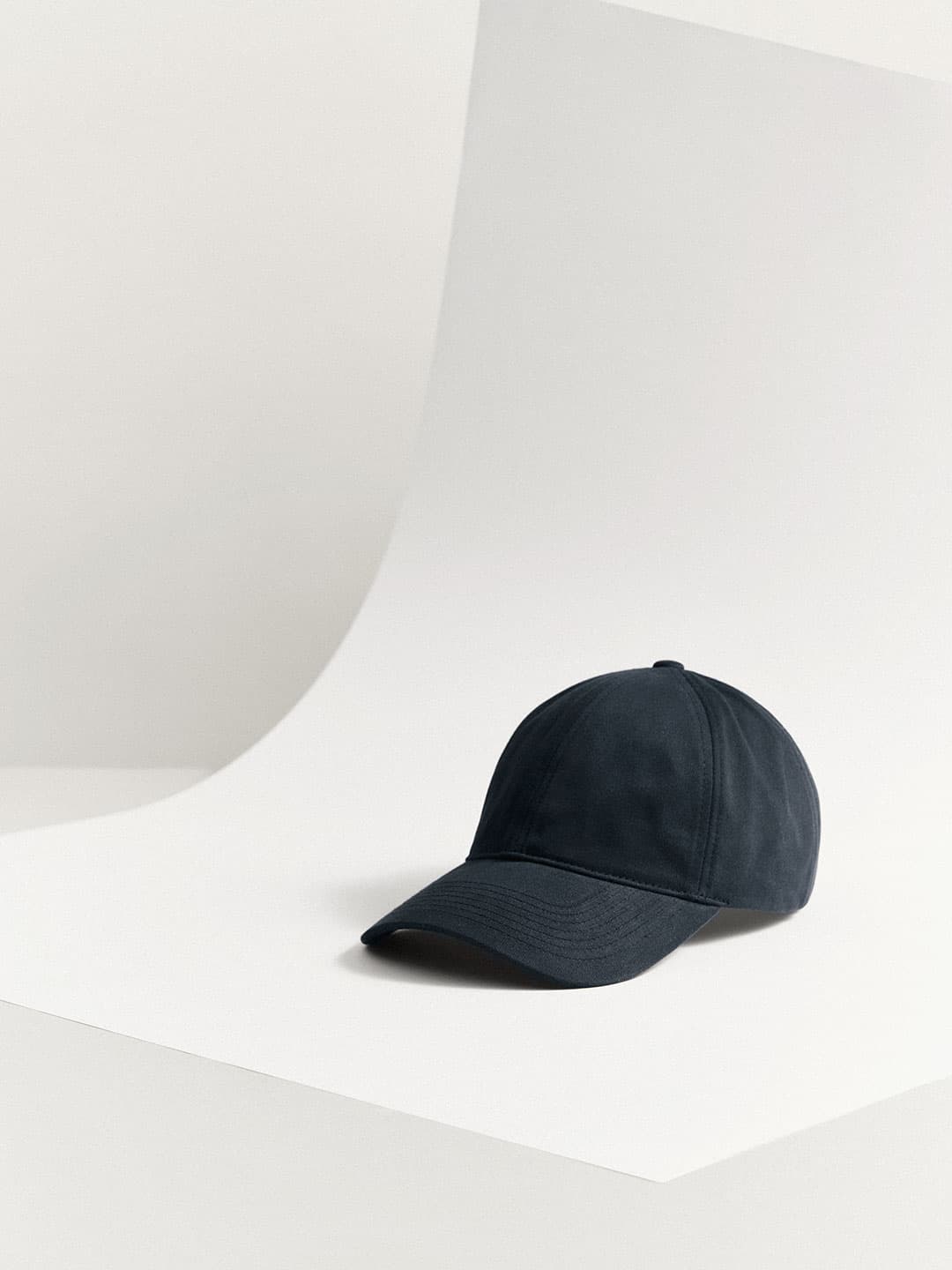 A black Cotton Twill baseball cap from Varsity Headwear is placed on a curved, light-colored surface in a minimalist setting.