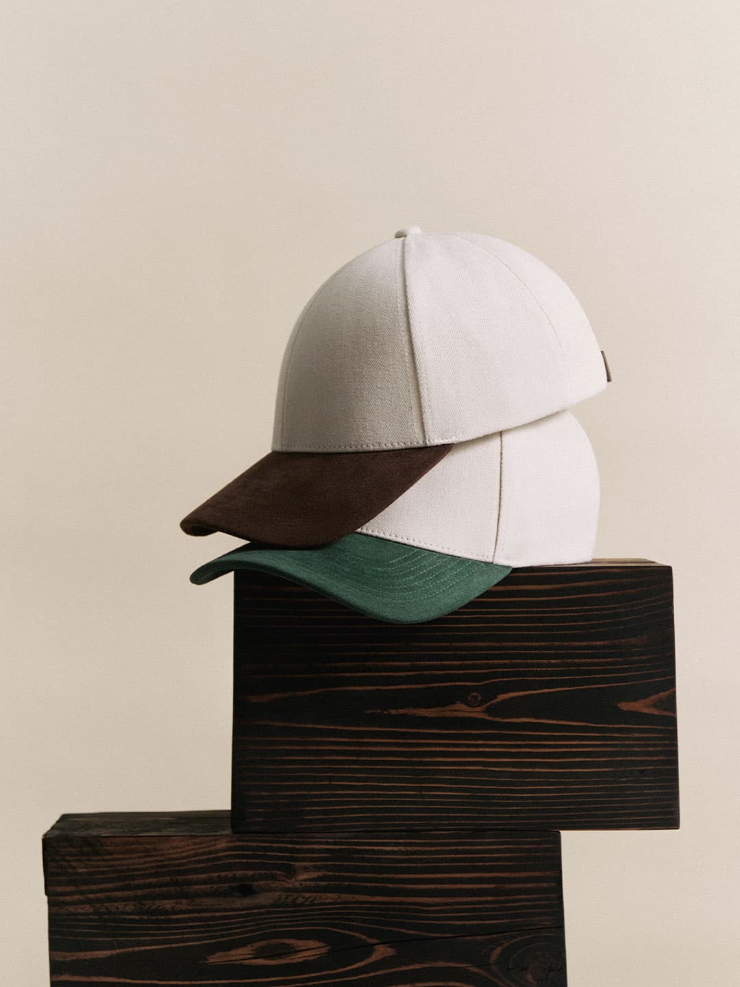 Two stacked baseball caps with beige crowns. One has a brown brim, and the other a green brim, set against a wooden surface and neutral background.
