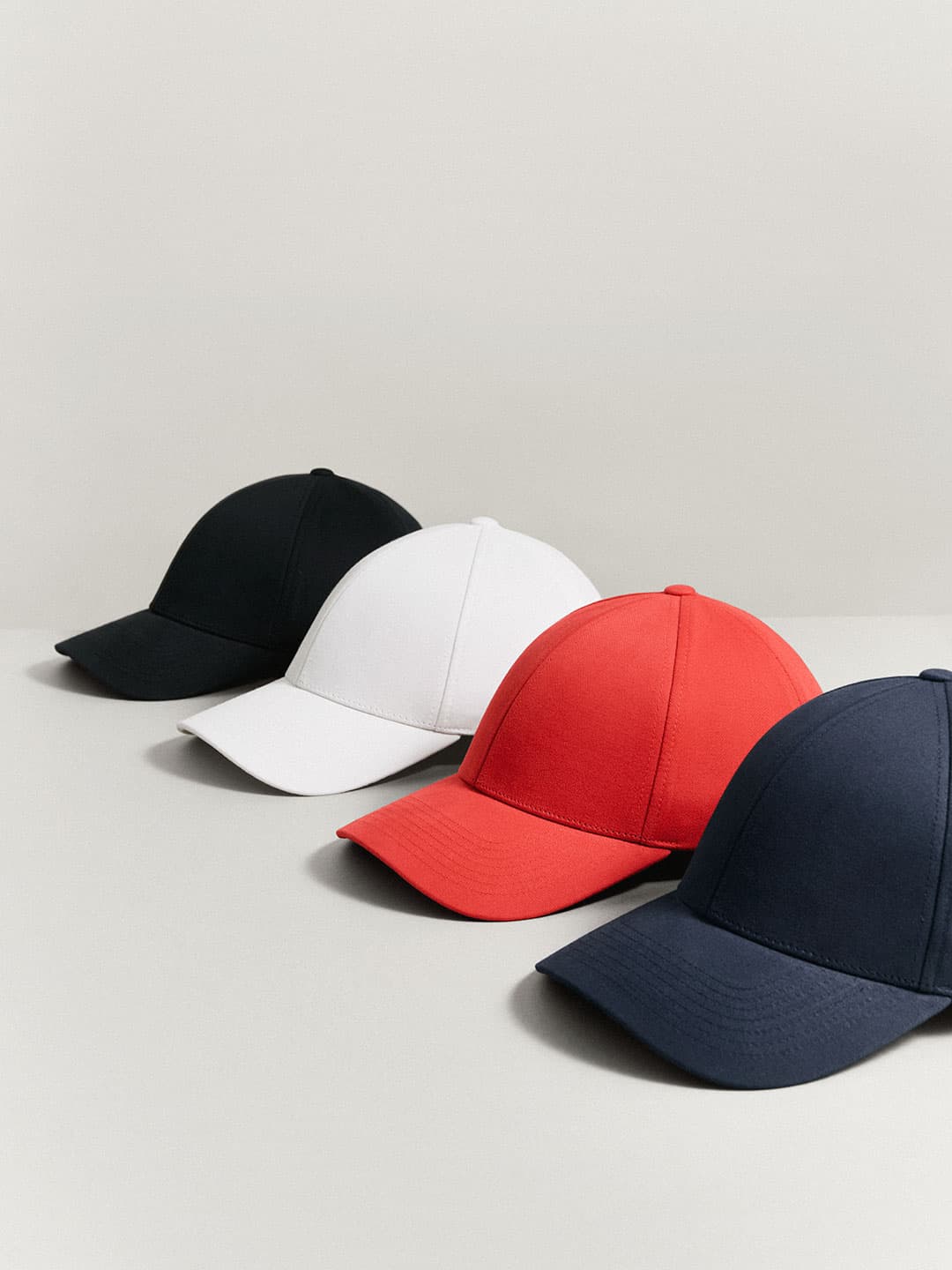 Four baseball caps in black, white, red, and navy blue in Twill Cotton from Varsity Headweare arranged in a row on a light gray surface.