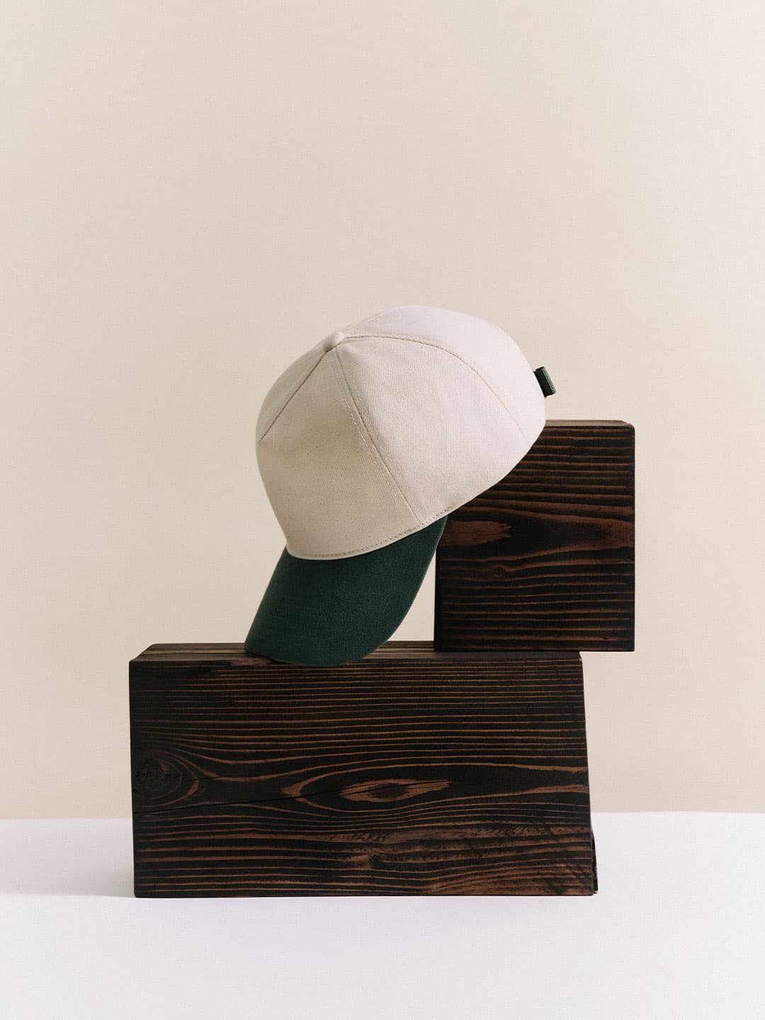 A beige denim cap with a green brim balanced on two stacked wooden blocks against a neutral background.