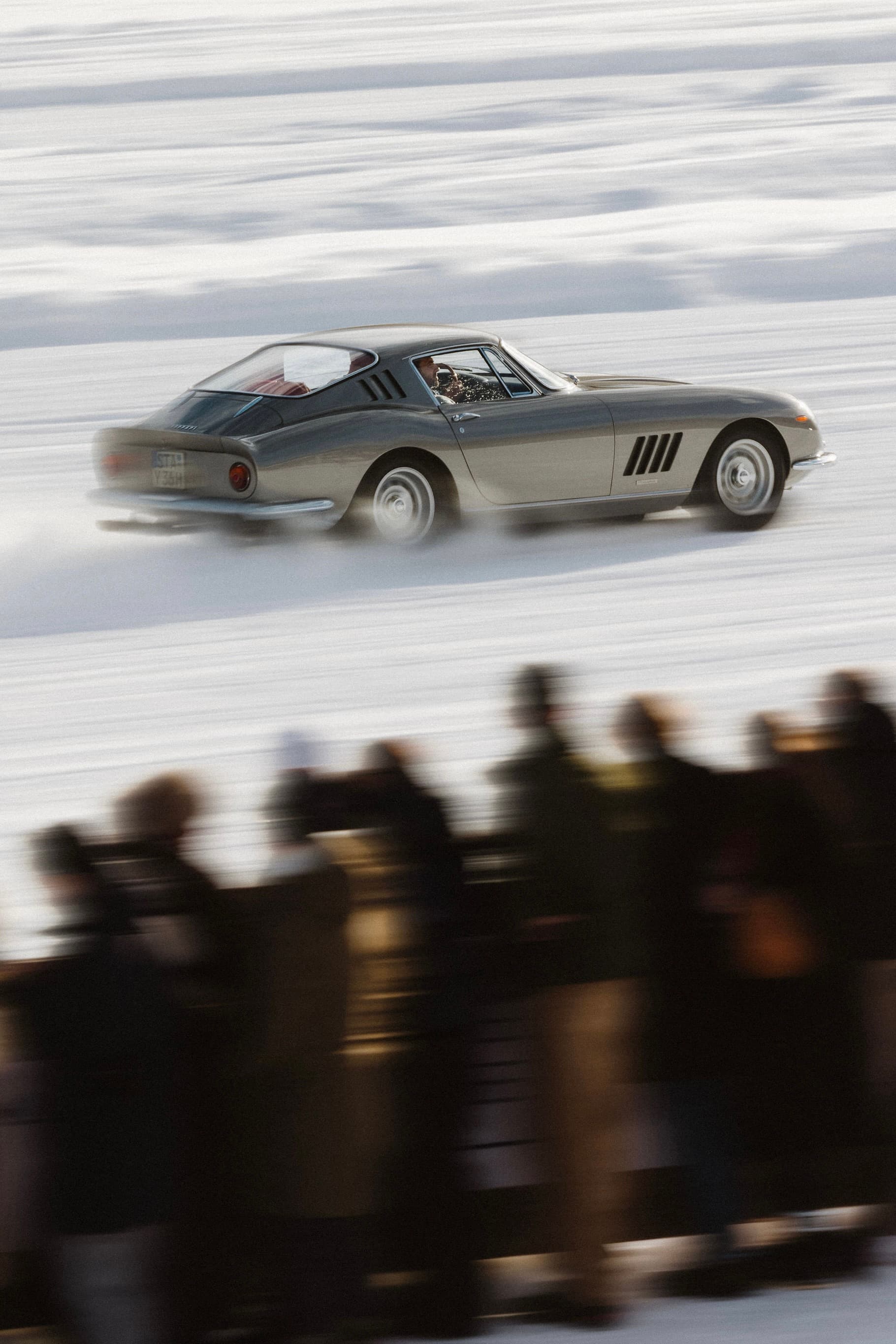 A classic car speeds across a snowy track with blurred spectators in the foreground, showcasing motion and excitement at The I.C.E in St. Moritz