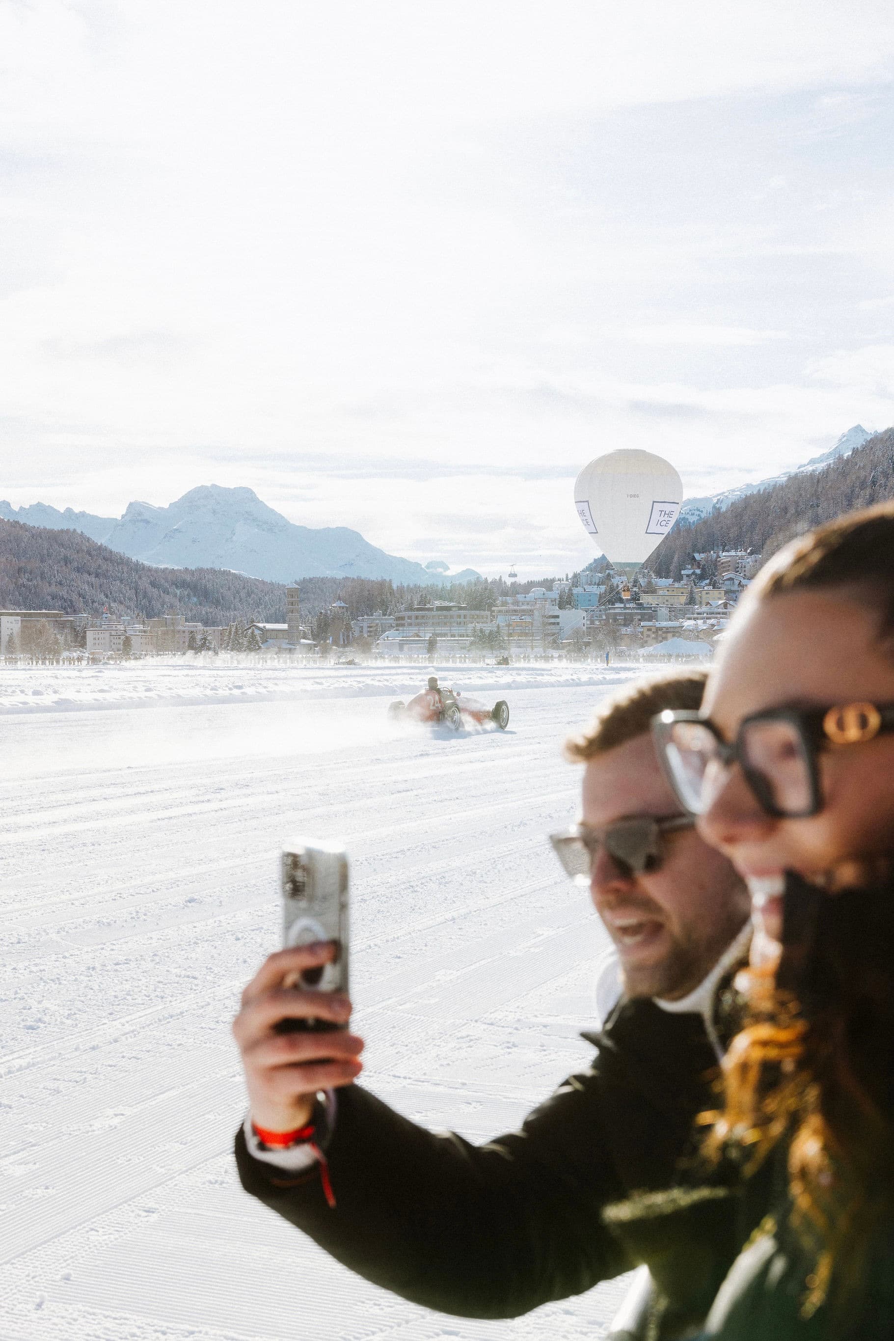 A man and a woman take a selfie in snowy terrain, with a vintage race car driving on snow and a hot air balloon in the background at The I.C.E in St. Moritz