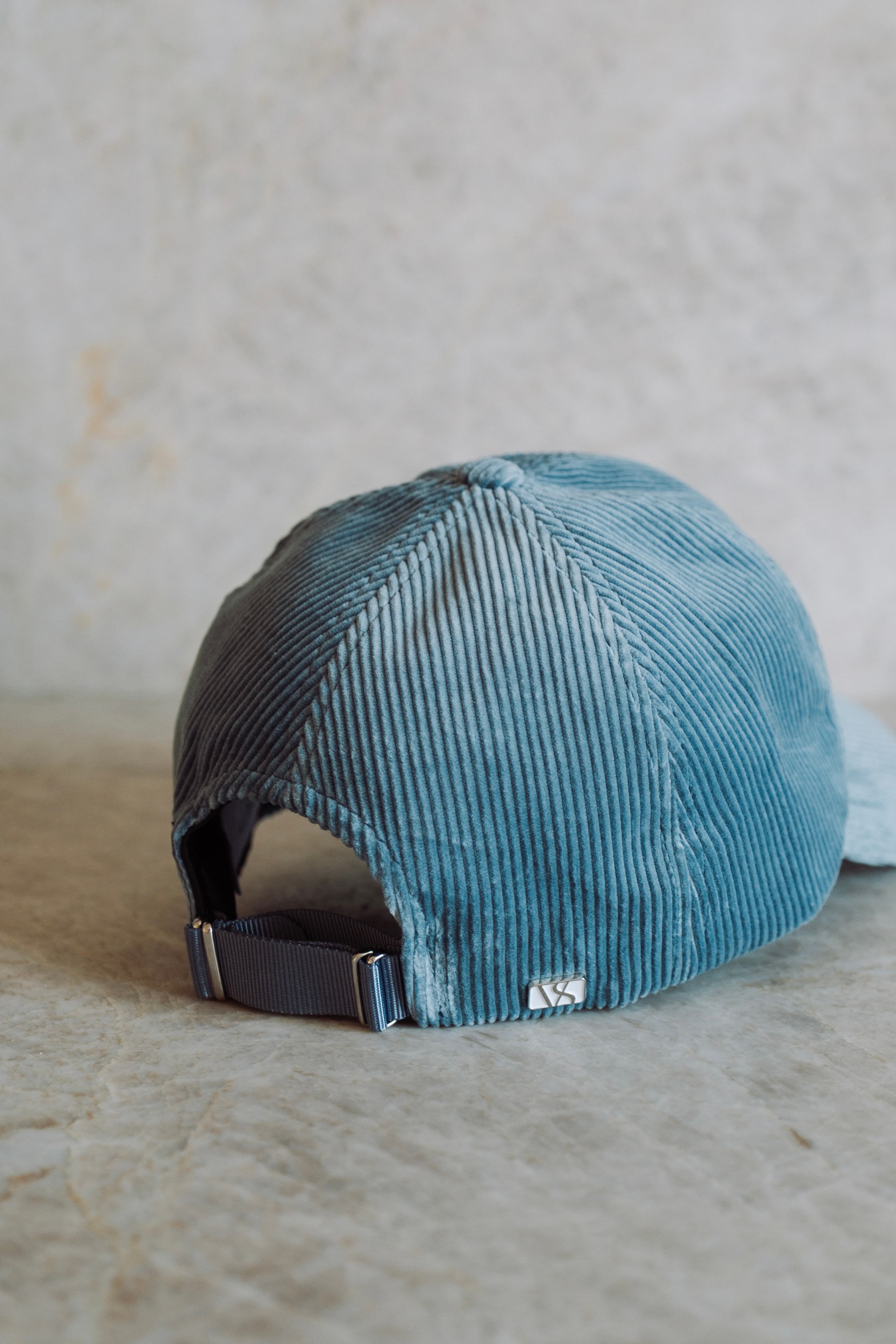 Blue corduroy baseball cap with an adjustable strap, displayed on a light stone background.