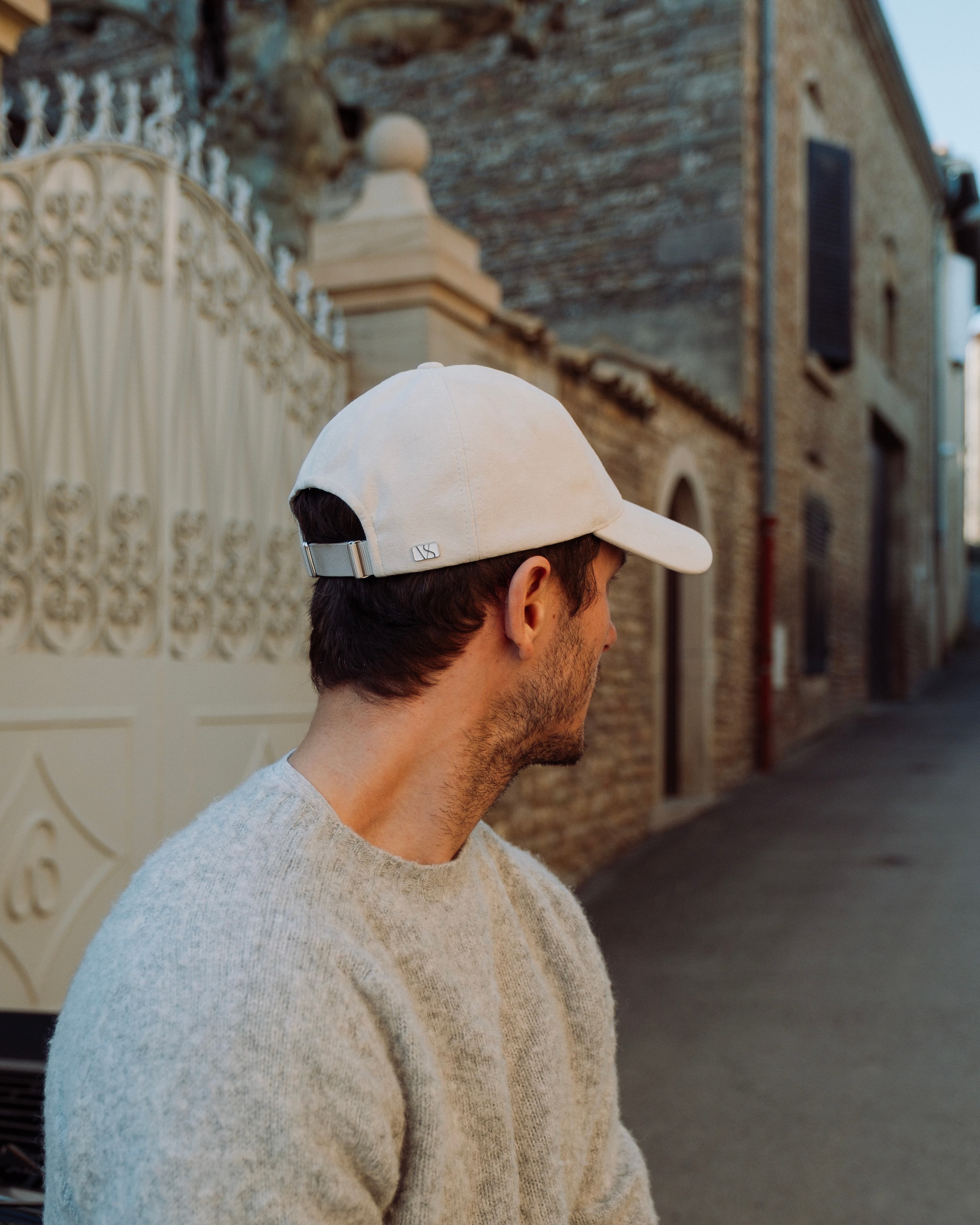 Man in a light sweater and white cap looks over his shoulder, standing near an ornate gate and brick building on a narrow street.
