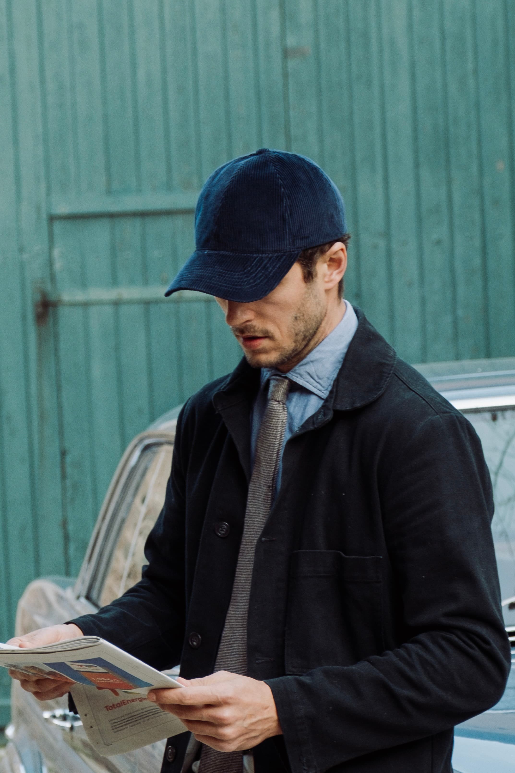 Hedi Sersoub Varsity Headwear ambassador wearing a navy corduroy cap from Varsity Headwear while reading the paper next to his vintage car